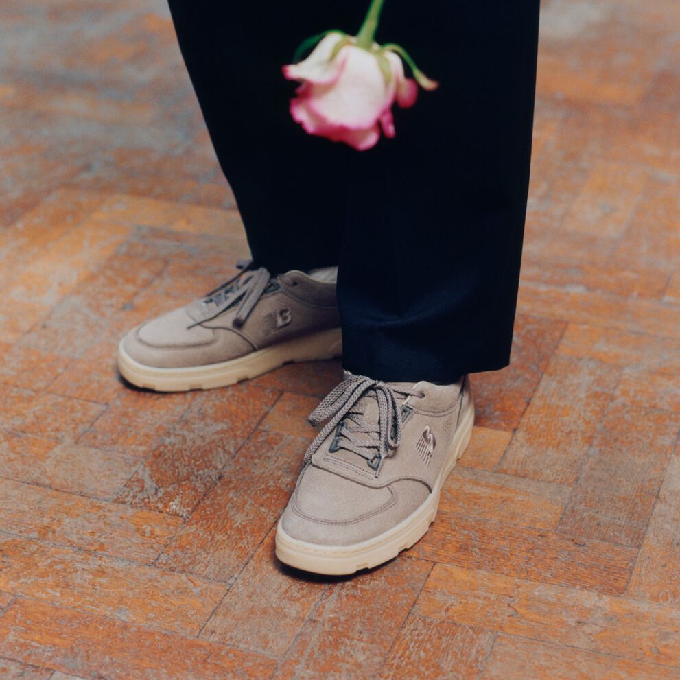 Knee down view of a man wearing a pair of New Balance Made in UK sneakers standing on a herringbone pattern floor with a white and pink rose being held in frame. 