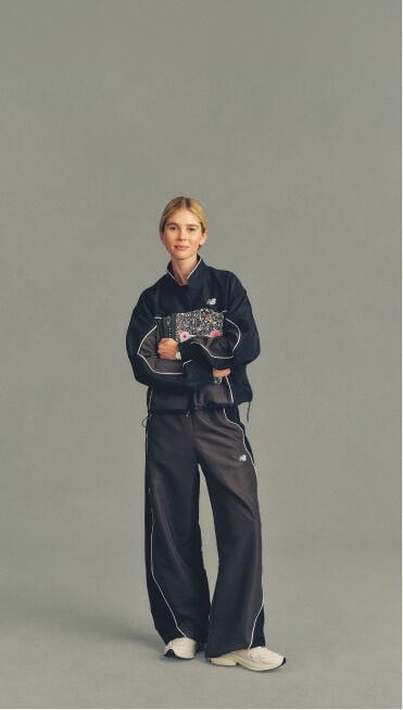 Full-body studio image of a person in a black New Balance tracksuit holding a patterned notebook against a neutral gray background.
