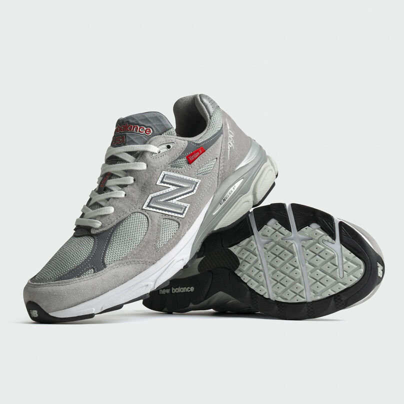 Pair of grey 990v3 sneakers stacked on each other in a grey studio environment.