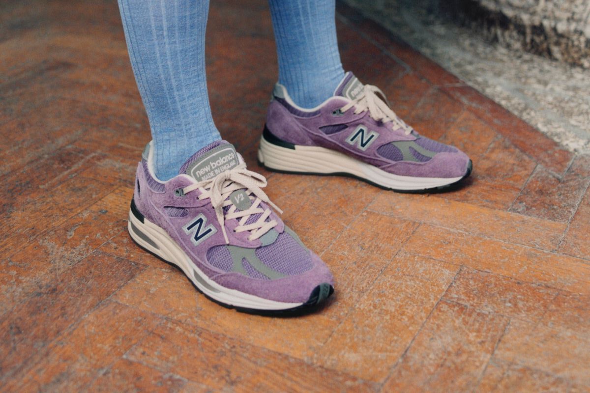 A pair of New Balance Made in UK sneakers standing on a wooden floor with textured green walls and a stone pillar in the background. 